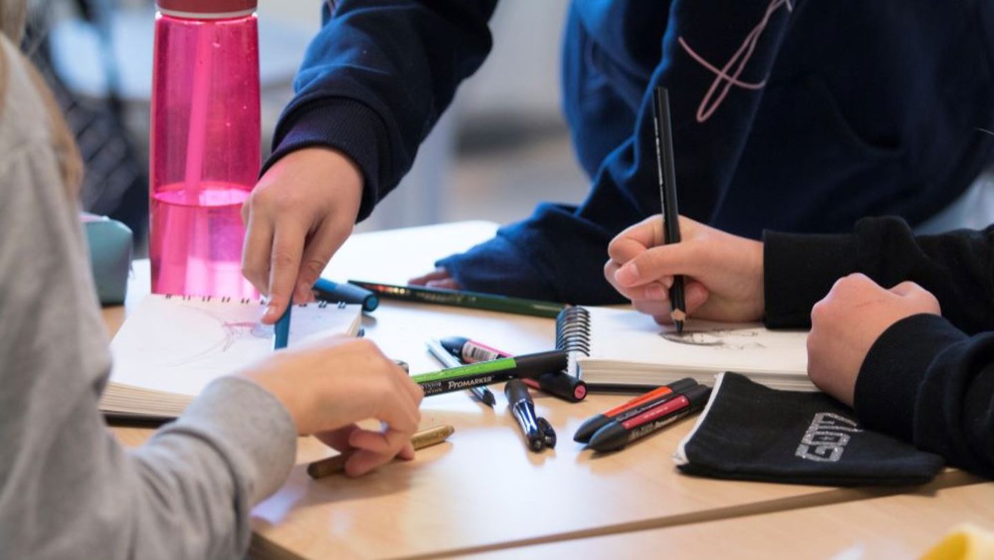 Bildet viser hendene til elever som sitter i et klasserom og jobber med oppgaver på et ark. 