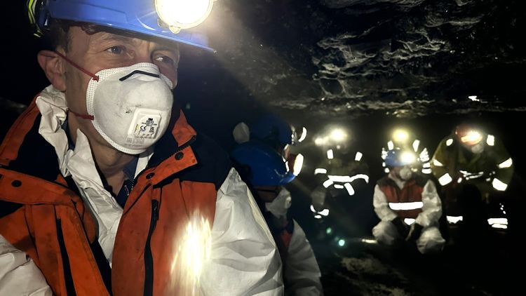 Mann i verneutstyr som hjelm, maske og dress ser i kamera mens flere gruvearbeidere sees i bakgrunnen