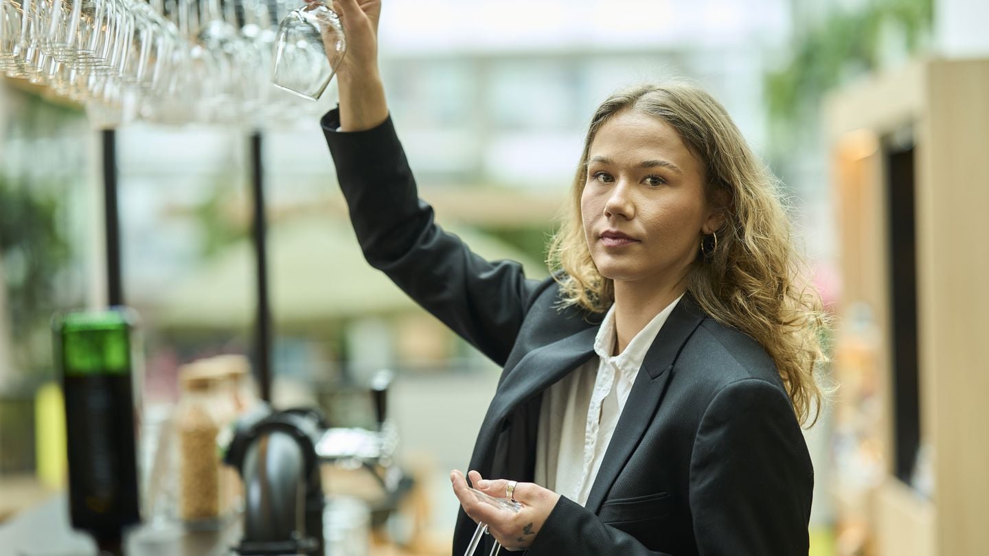  En kvinnelig bartender med blazer rekker opp hånden mot en hylle med hengende vinglass. I bakgrunnen står flasker, kaffemaskin og annet utstyr som brukes til servering.